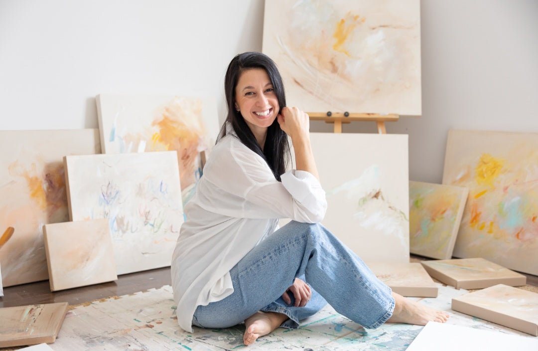 Artiste assise entourée de toiles abstraites aux couleurs douces dans un atelier lumineux
